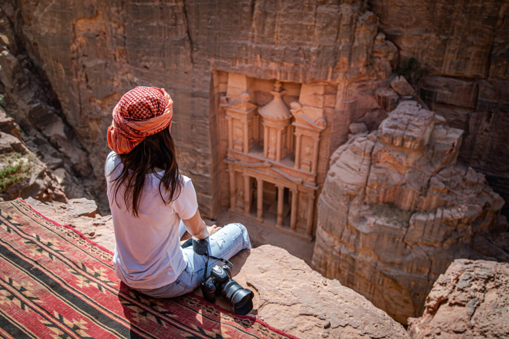 Asian woman traveler sitting on carpet viewpoint in Petra ancient city looking at the Treasury or Al-khazneh, famous travel destination of Jordan and one of seven wonders. UNESCO World Heritage site.