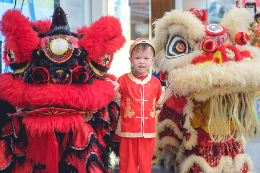 Cute smiling little 2 - 3 years old Asian toddler baby boy child in traditional red Chinese costume celebrating Chinese New Year with Chinese lion dance in Bangkok, Lunar New Year celebration concept