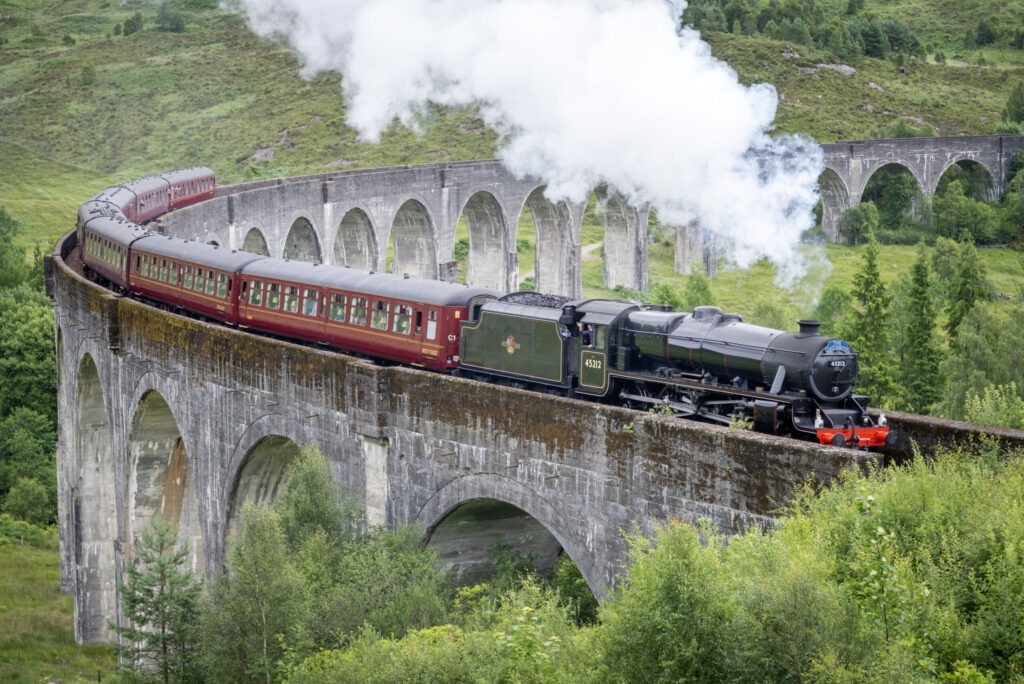 Glenfinnan,Inverness-shire,Scottish Highlands-July 21 2022:The train driver waves and lets off steam to onlookers,as the iconic train,shown in Harry Potter films,crosses the tall viaduct,mid summer.